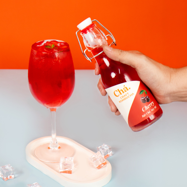 Một bàn tay cầm chai Chá. Cherry bên ly kombucha đỏ, trên nền cam và mặt bàn xanh nhạt.